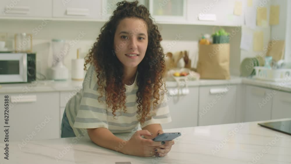 Medium shot of young curly haired girl leaning on kitchen table, holding smartphone and posing for camera during day at home Medium shot of young curly haired girl leaning on kitchen table, holding smartphone and posing for camera during day at home