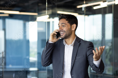 Successful joyful businessman talking on the phone, mature hispanic man standing inside office and talking with clients, telling colleagues good successful news.