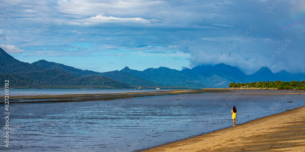 Fototapeta premium pretty girl in yellow dress admiring the view of hinchinbrook island from the beach in cardwell, north queensland, australia; tropical beach with palm trees and famous island in the background