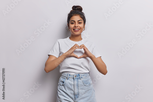 Good looking Indian woman radiates love her hands forming heartfelt gesture reminding us of power of love and compassion smiles broadly dressed in casual t shirt and jeans isolatd on white background