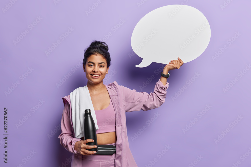 Φωτογραφία Positive sporty woman holds communication bubble and bottle of water smiles gladfully poses with towel on shoulder suggests to write your ideas here isolated over purple background