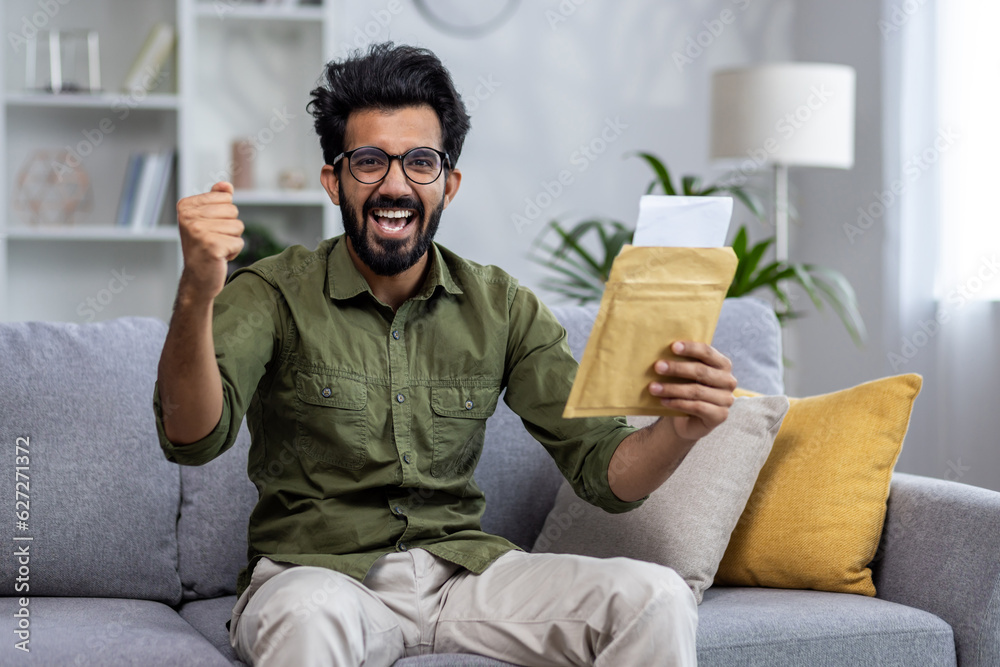 Portrait of successful satisfied man at home, hispanic man holding message envelope in hands and happy celebrating victory, looking joyfully at camera sitting on sofa in living room.