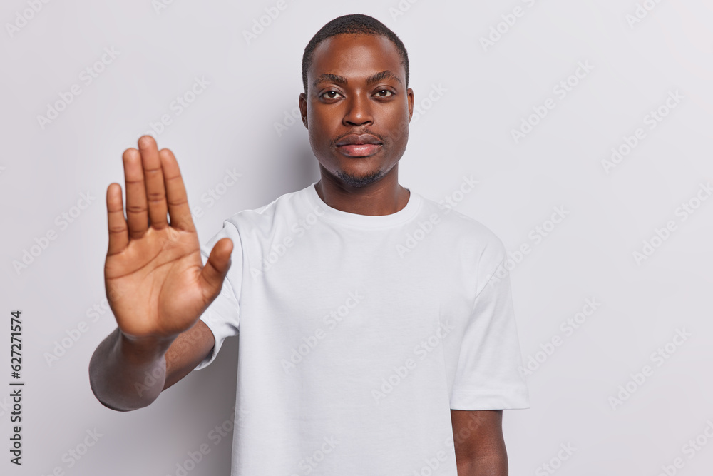 Concept of prohibition and refusal. Dark skinned man raises palm in stop gesture clearly communicates his objection and says no asserting boundaries wears casual t shirt isolated over white background Concept of prohibition and refusal. Dark skinned man raises palm in stop gesture clearly communicates his objection and says no asserting boundaries wears casual t shirt isolated over white background