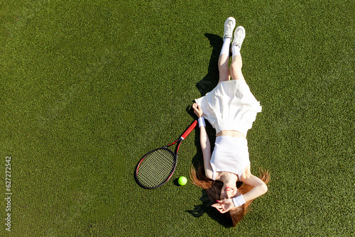 young girl tennis player in white sports uniform with tennis racket lies and rests on green court, top view