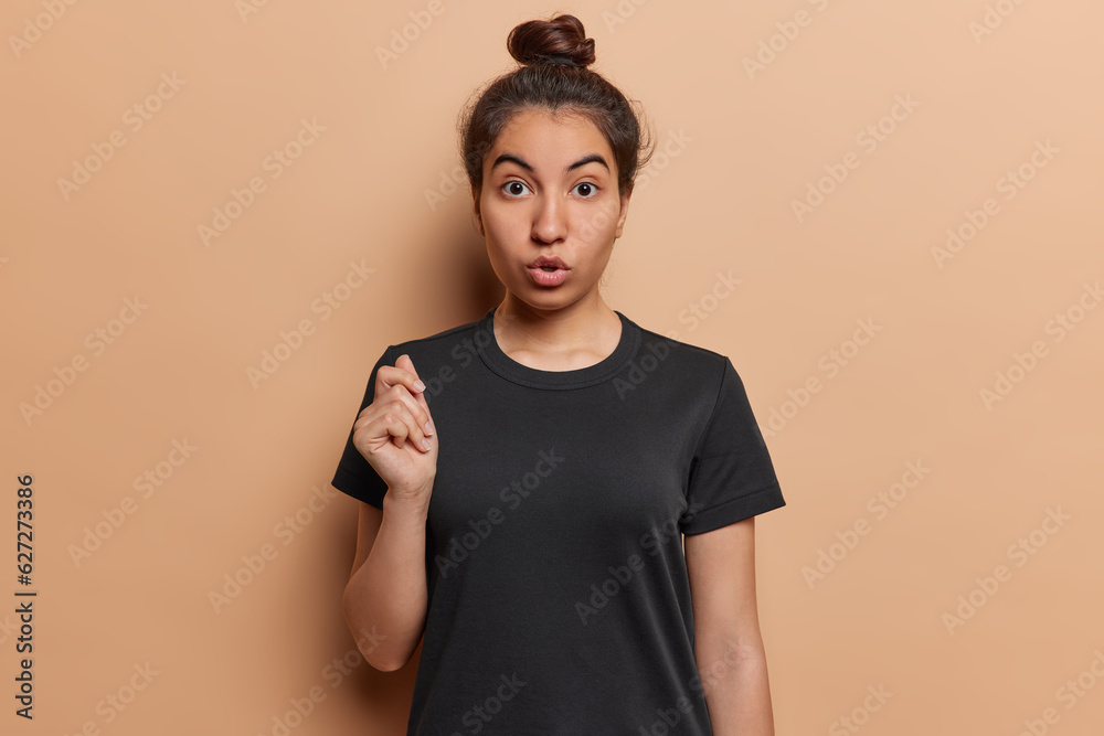 Φωτογραφία Waist up shot of impressed Iranian girl with hair bun reacts to something shocking holds breath from amazement dressed in casual black t shirt isolated over brown background