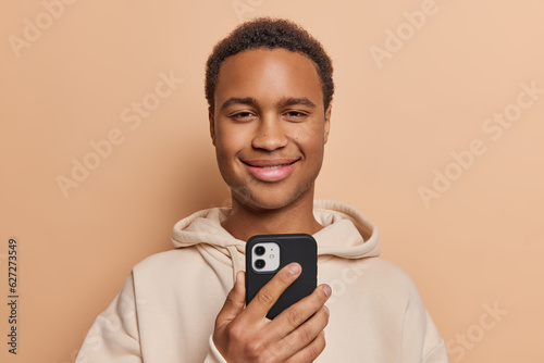 Portrait of handsome dark skinned man dressed in casual hoodie uses smartphone for chatting onlie sends and gets messages isolated over brown background. People and online communication concept photo