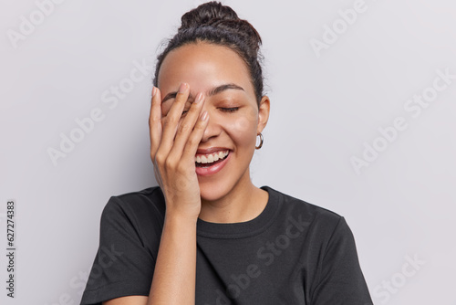 Studio shot of overjoyed Latin woman makes face palm laughs joyfully keeps eyes closed expresses positive emotions wears casual black t shirt isolated on white background. People and happiness concept