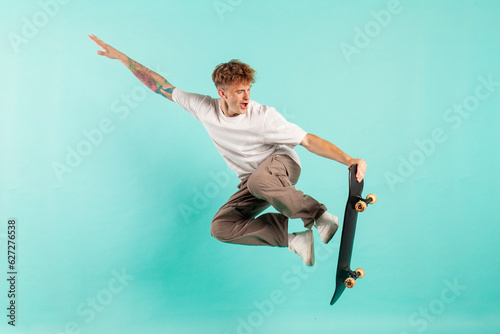 young crazy guy rides skateboard and jumps on blue isolated background, hipster in sunglasses flies