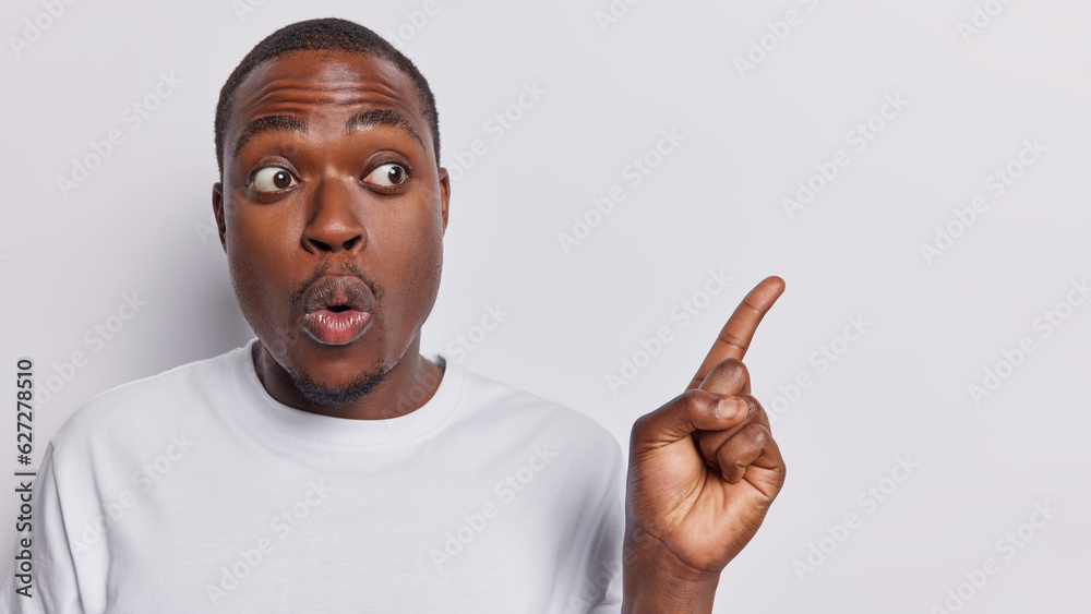 Φωτογραφία Photo of impressed dark skinned adult man pointing at copy space for advertisement or text showing great deal or offer keeps mouth opened gives recommendation poses against white background