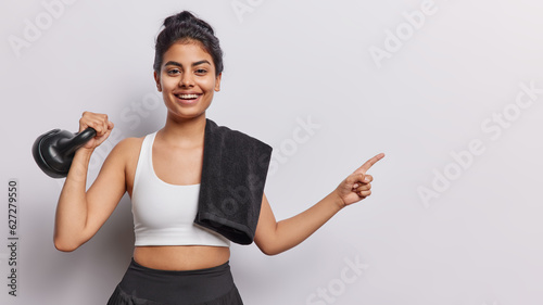 Waist up shot positive sporty woman with dark hair lifts weight points index finger at copy space for your advertising content dressed in sportswear towel on shoulder isolated over white background