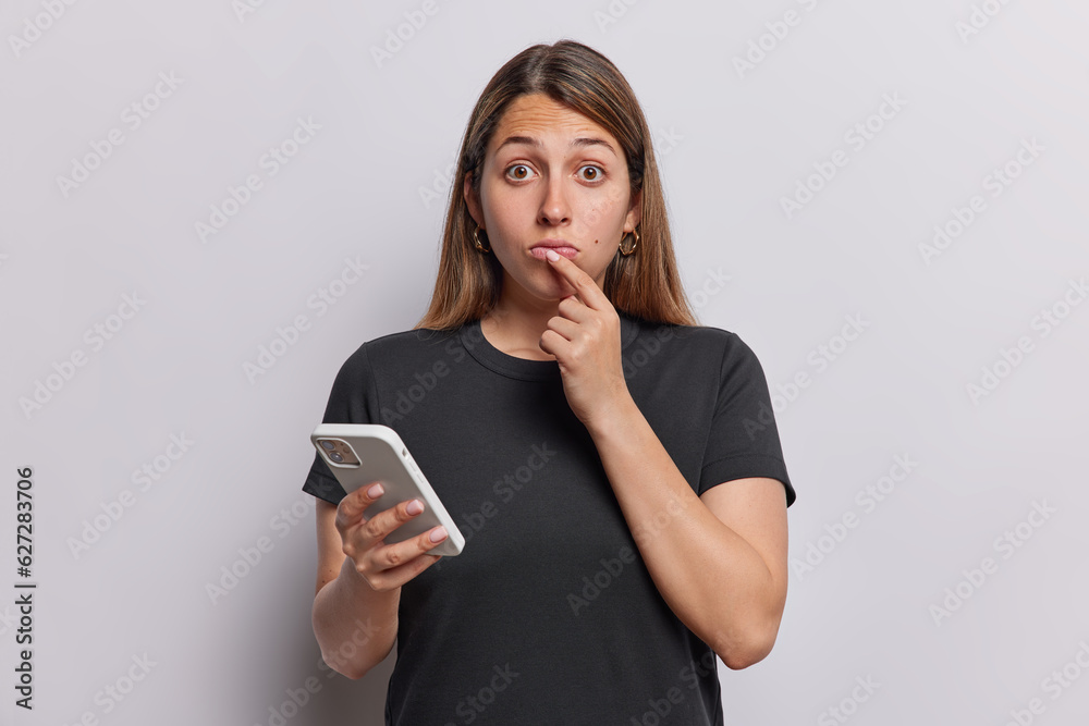 Surprise long haired woman keeps finger on lips reacts to something shocking holds mobile phone in hand wears casual black t shirt isolated over white background. People and technology concept Surprise long haired woman keeps finger on lips reacts to something shocking holds mobile phone in hand wears casual black t shirt isolated over white background. People and technology concept
