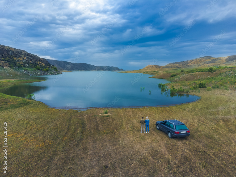 Drone view of a lake, a car, a man and a woman looking into the distance among small mountains. Beautiful landscape from a drone. Concept of rest, travel.