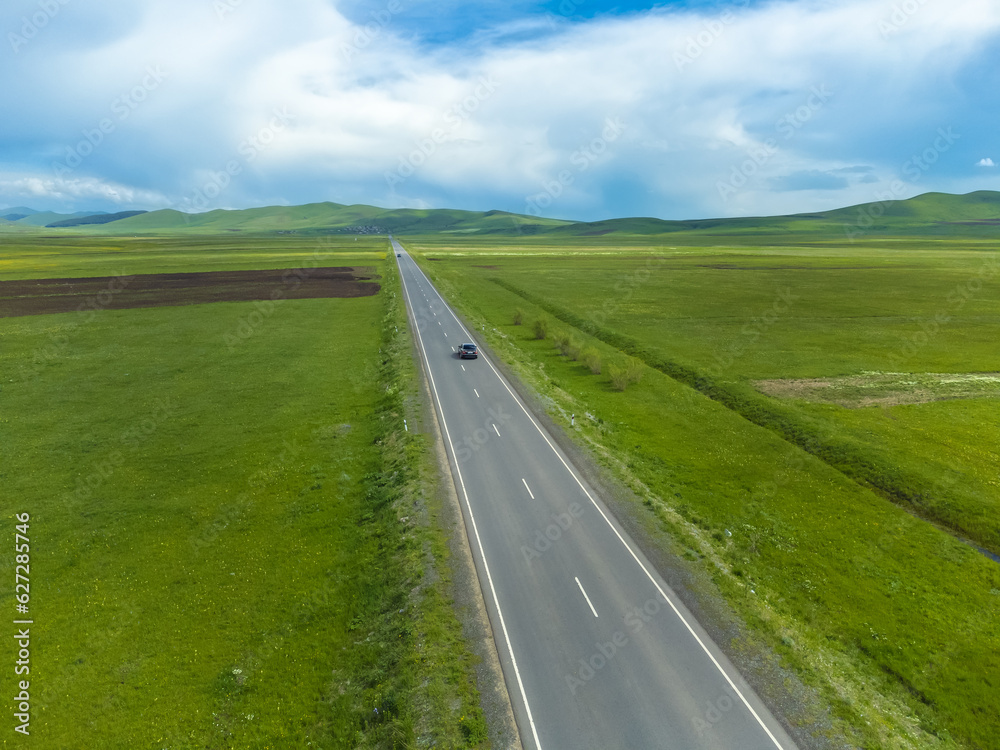 Black passenger car is driving along an asphalt road along green fields, mountains on a sunny day. Photographing from a drone. Concept of travel