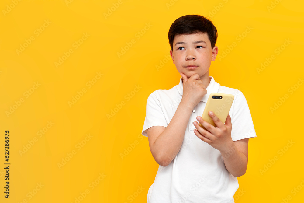 pensive asian little boy of ten years old in white t-shirt uses smartphone and thinks on yellow background pensive asian little boy of ten years old in white t-shirt uses smartphone and thinks on yellow background
