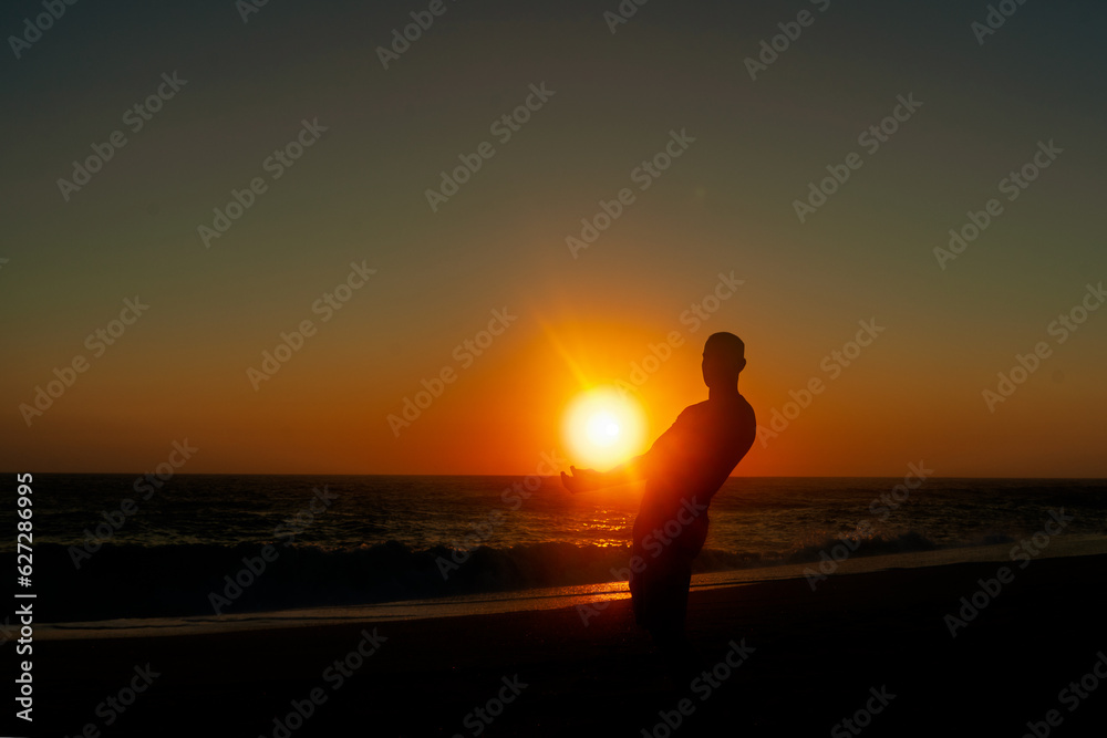 Krajobraz morski, sylwetka człowieka podnosząca słońce. Zachód słońca na plaży.  - obrazy, fototapety, plakaty 