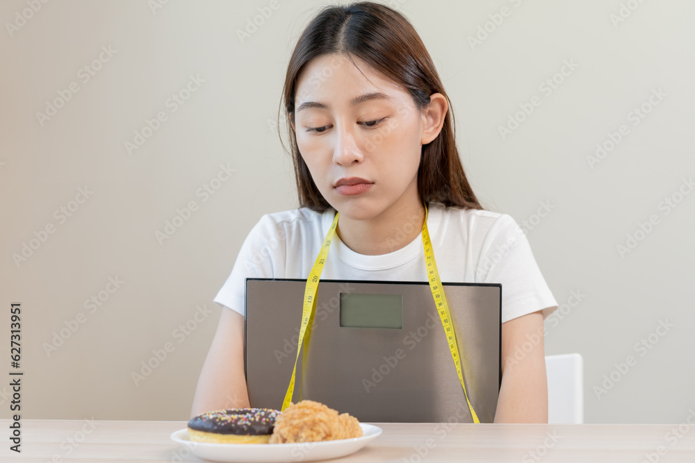Diet, attractive asian young woman, girl restrained to eat doughnut, bakery and fried chicken, fast food to lose, loss weight, hugging weight scales on table at home. Passion, temptation when hungry. Diet, attractive asian young woman, girl restrained to eat doughnut, bakery and fried chicken, fast food to lose, loss weight, hugging weight scales on table at home. Passion, temptation when hungry.