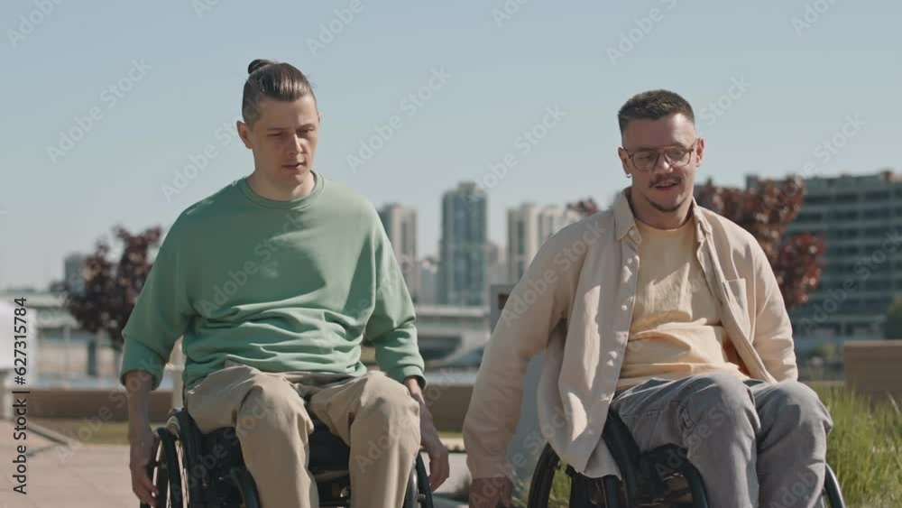 Medium shot of two Caucasian male friends chatting while moving in wheelchairs along city embankment on warm sunny day Medium shot of two Caucasian male friends chatting while moving in wheelchairs along city embankment on warm sunny day