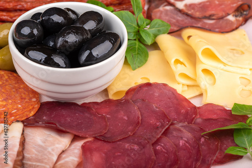Close-up of olives in a white bowl, sliced prosciutto and cheese, green basil.