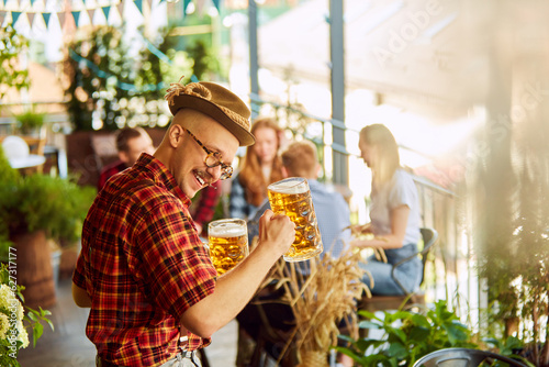 Close-up of young man in checkered shirt and fedora hat drinking beer. Blurred people in background. Sunny day. Concept of oktoberfest, traditional taste, friendship, leisure time, enjoyment © master1305