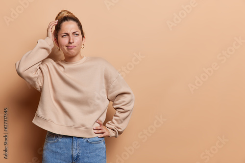 Studio shot of pensive young woman scratches head stands thoughtful considers something dressed in casual pullover and jeans isolated over brown background copy space for your advertising content