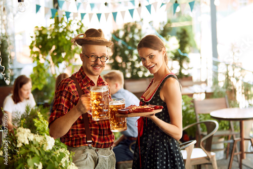 Man and woman in traditional clothes standing with lager beer and appetizers at pub. Blurred people on background. Concept of oktoberfest, traditional taste, friendship, leisure time, enjoyment
