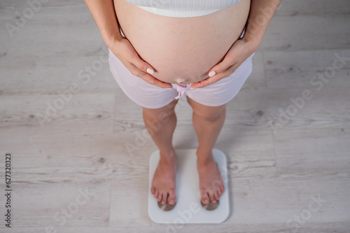 Top view of a pregnant woman in home clothes standing on an electronic scale and holding her hands on her tummy. 