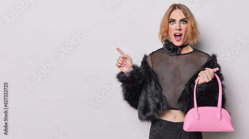 Photo of shocked transgener woman with vivid makeup dressed in outerwear points finger aside on copy space demonstrates something awesome isolated over white background showcases his unique style
