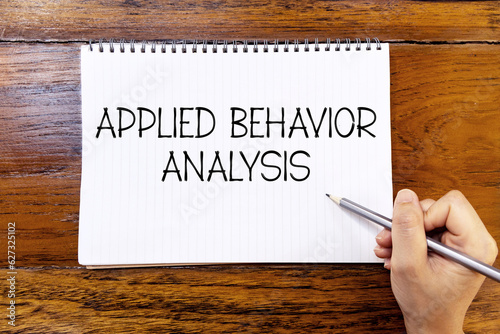 Applied behavior analysis handwriting text on blank notebook paper on wooden table with hand holding pencil. Business concept and legal concept about applied behavior analysis.