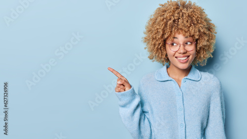 Horizontal shot of cheerful blonde curly young woman points index finger aside drawing attention to copy space demonstrates advertisement dressed in casual jumper isolated over blue background