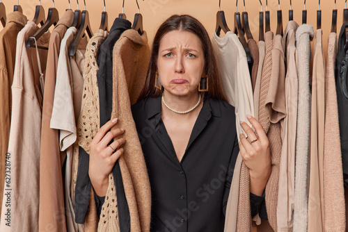 Horizontal shot of displeased European woman purses lips stands near display rack filled with hangers immerse herself in world of fashion poses around clothes in wardrobe. Beige color prevails