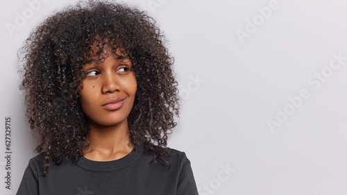 Studio shot of curly haired woman concentrated aside purses lips feels dissatisfied wears casual black t shirt being deep in thoughts isolated on white background copy space for your promotional text