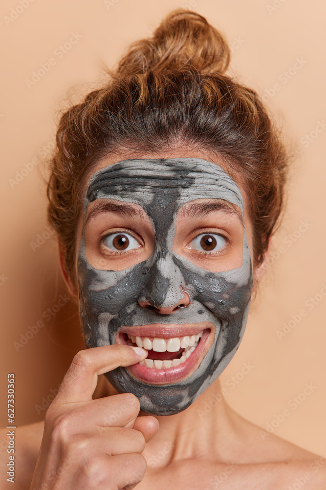 Vertical shot of young woman engages in blissful beauty ritual at home bites finger stands shirtless undergoes skin care procedures isolated over beige background indulging in the soothing treatments Vertical shot of young woman engages in blissful beauty ritual at home bites finger stands shirtless undergoes skin care procedures isolated over beige background indulging in the soothing treatments