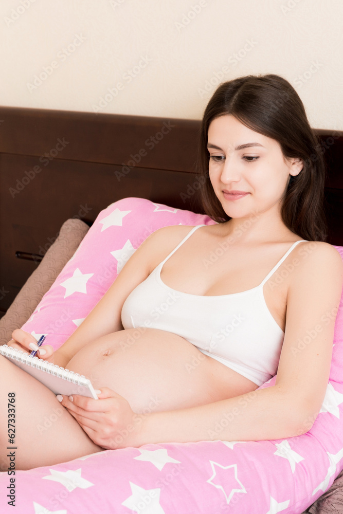 Obraz premium Pregnant woman writing baby names list on sofa, closeup. Pregnant mom writes in a notebook and to-do list shopping