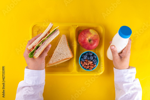 Healthy school meal, children packed lunch box with fruit, berry, nuts and sandwich with vegetables. Kids diet snack food with education school supplies, bright blue background top view copy space