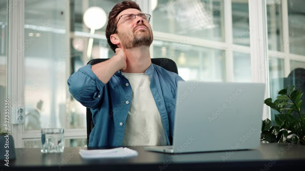 Pain in neck. Closeup of bearded man in office workplace, man experiences severe pain in neck, touches his neck with hand and makes constrained head movements. Hard work at a table with a laptop.