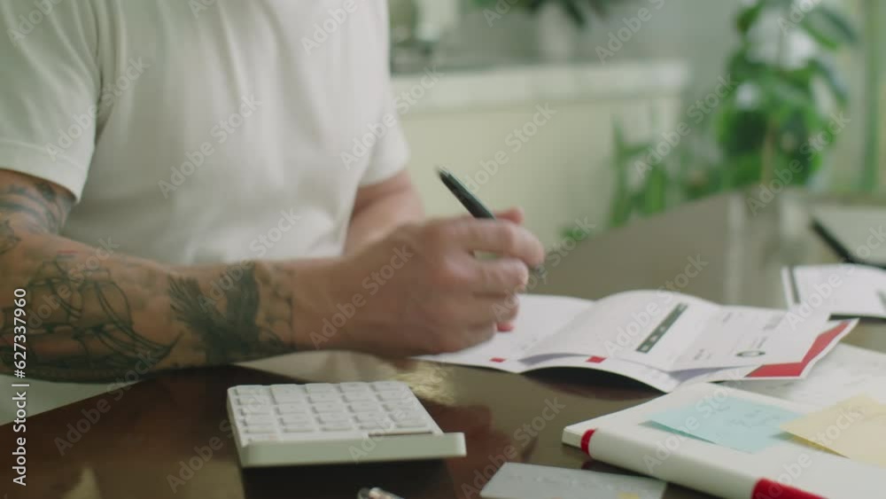 Medium close up view of young man checking bills, counting with calculator and writing down expenses while managing his finances at home Medium close up view of young man checking bills, counting with calculator and writing down expenses while managing his finances at home
