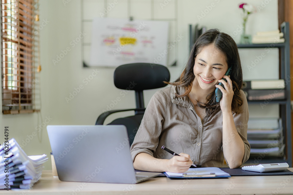 Asian businesswoman using smartphone talking to business client and doing math on wooden table working with laptop in office, tax, accounting, finance concept Asian businesswoman using smartphone talking to business client and doing math on wooden table working with laptop in office, tax, accounting, finance concept