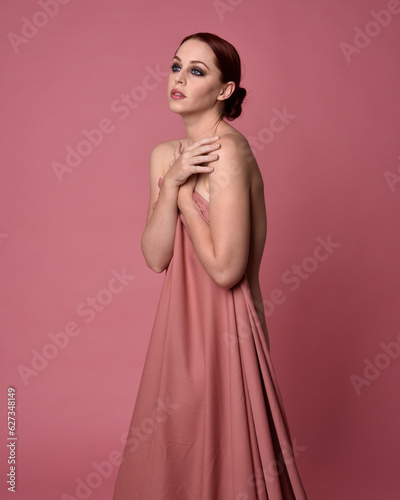 Portait of beautiful woman wearing gown of draped silk fabric, standing pose. Isolated on pink studio background.
