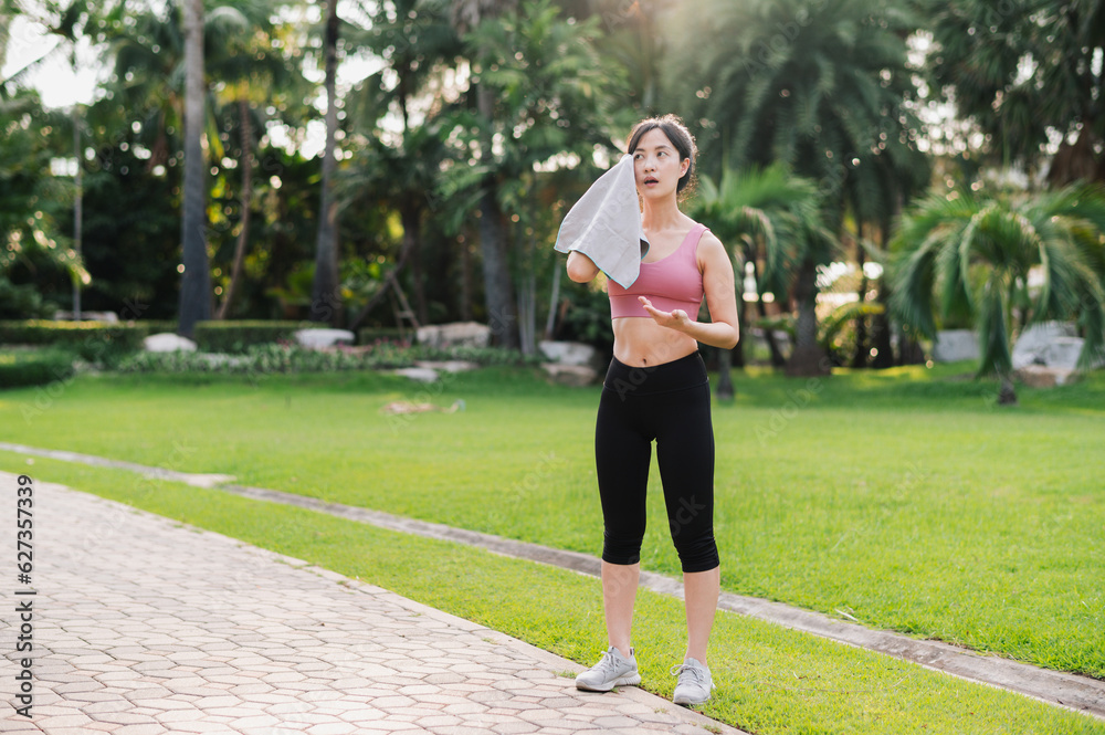 Embrace the beauty of a healthy lifestyle as 30s Asian woman in pink sportswear wipes her cheek after a morning run in the public park. Achieve fitness goals and experience a sense of accomplishment.