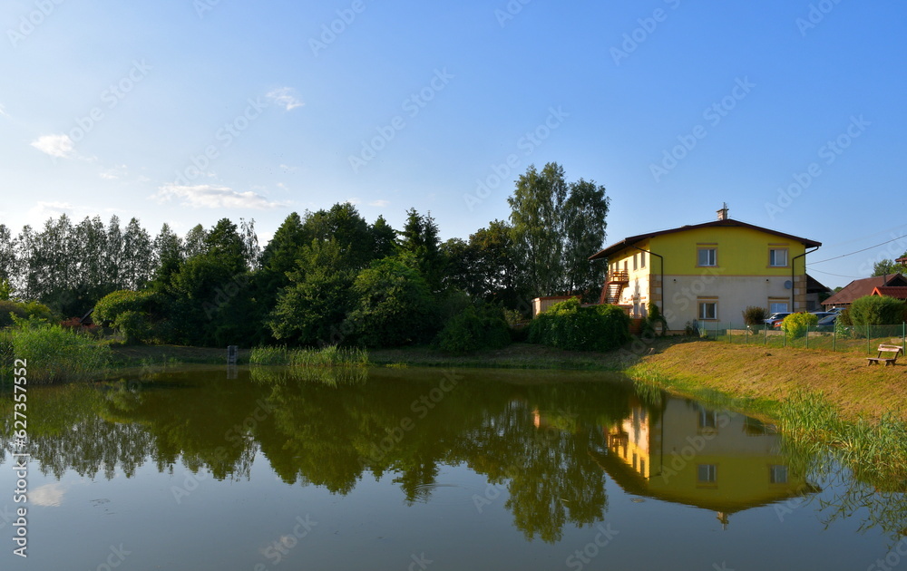 A view of a rural home, house, shelter, or shack located next to a dense forest or moor located next to the coast or bank of a shallow yet vast river, lake or pond seen on a sunny summer day