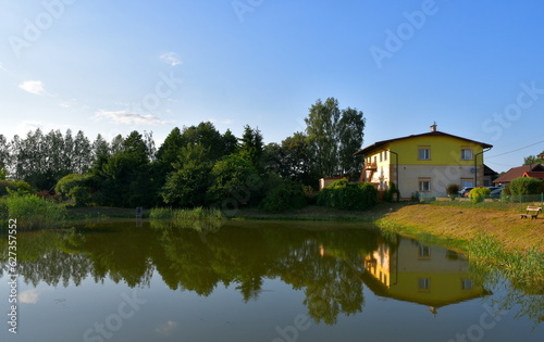 A view of a rural home, house, shelter, or shack located next to a dense forest or moor located next to the coast or bank of a shallow yet vast river, lake or pond seen on a sunny summer day