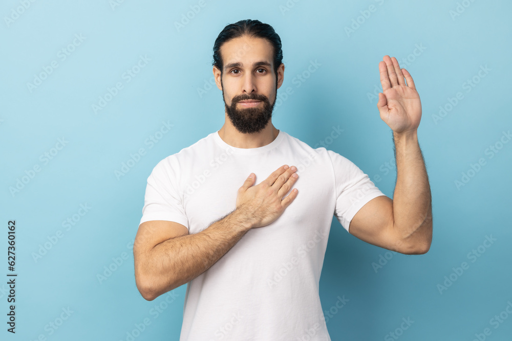 I swear. Conscious honest man with beard in white T-shirt raising one hand and holding on chest another, swearing in honesty and devotion, patriotism. Indoor studio shot isolated on blue background. I swear. Conscious honest man with beard in white T-shirt raising one hand and holding on chest another, swearing in honesty and devotion, patriotism. Indoor studio shot isolated on blue background.