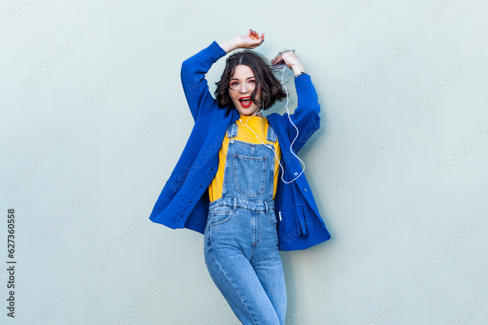 Portrait of happy cheerful hipster woman in denim overalls, yellow T-shirt and blue jacket, listening to music, dancing with raised hands. Indoor studio shot isolated on light green background. Portrait of happy cheerful hipster woman in denim overalls, yellow T-shirt and blue jacket, listening to music, dancing with raised hands. Indoor studio shot isolated on light green background.