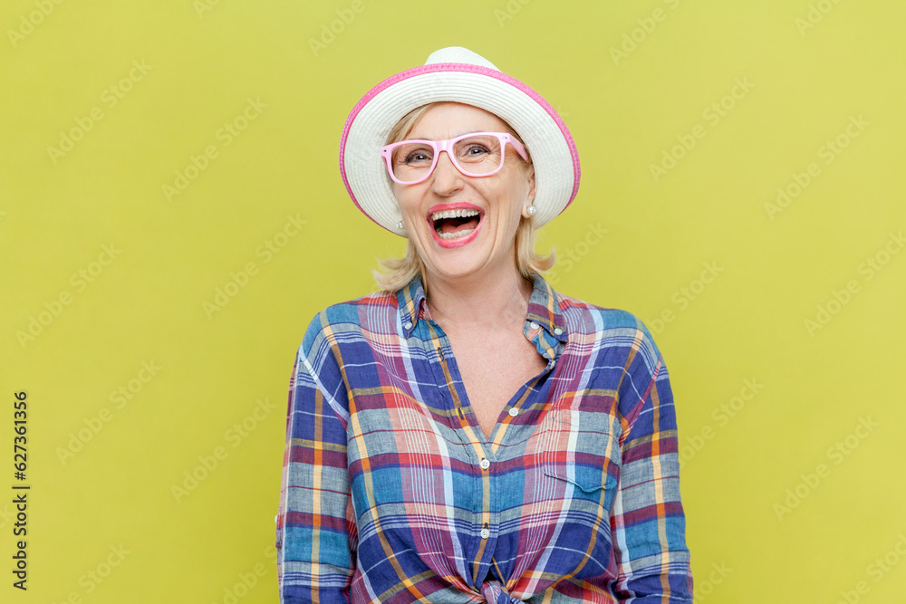 Portrait of laughing senior woman wearing checkered shirt, hat and eyeglasses being very glad, smiling with broad smile showing her perfect mood. Indoor studio shot isolated on yellow background. Portrait of laughing senior woman wearing checkered shirt, hat and eyeglasses being very glad, smiling with broad smile showing her perfect mood. Indoor studio shot isolated on yellow background.