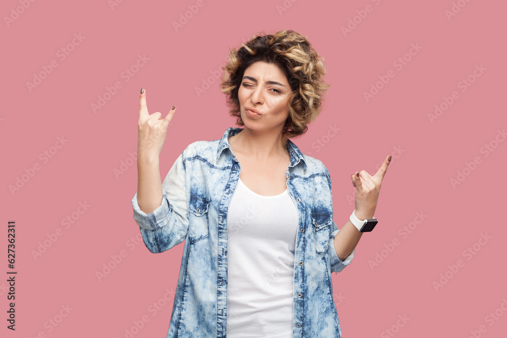 Portrait of cool festive woman with curly hairstyle wearing blue shirt standing and showing rock and roll gesture, enjoying concert. Indoor studio shot isolated on pink background. Portrait of cool festive woman with curly hairstyle wearing blue shirt standing and showing rock and roll gesture, enjoying concert. Indoor studio shot isolated on pink background.