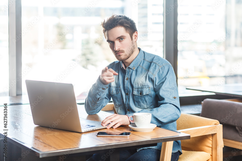 Portrait of strict young man freelancer in blue jeans shirt working on laptop and pointing finger to camera, saying you must to finish project. Indoor shot near big window, cafe background. Portrait of strict young man freelancer in blue jeans shirt working on laptop and pointing finger to camera, saying you must to finish project. Indoor shot near big window, cafe background.