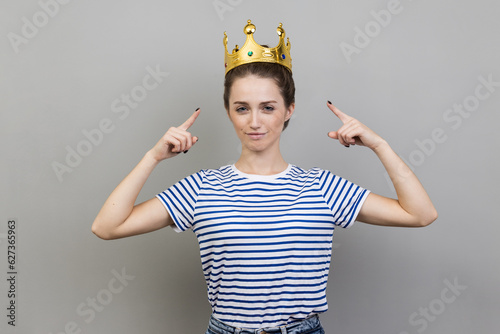 Portrait of confident woman wearing striped T-shirt, pointing at golden crown on her head, looking with arrogance and confidence, privileged status. Indoor studio shot isolated on gray background.