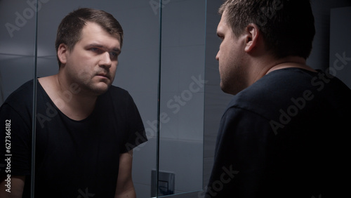 Young concentrated man feeling emotional pressure looking in his reflection in mirror