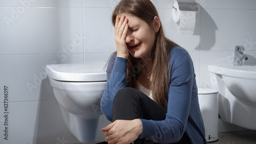 Crying young woman sitting on floor in toilet. Concept of depression, home violence, suicide, stress, loneliness and frustration.