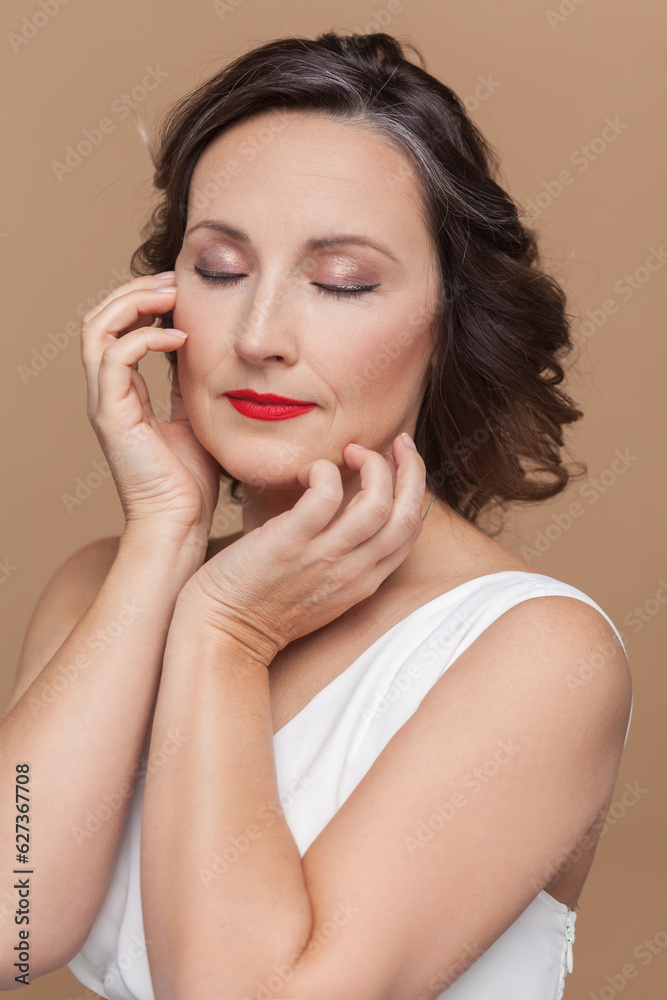 Closeup portrait of calm attractive beautiful middle aged woman with wavy hair and makeup, keeps eyes closed, wearing white dress. Indoor studio shot isolated on light brown background.